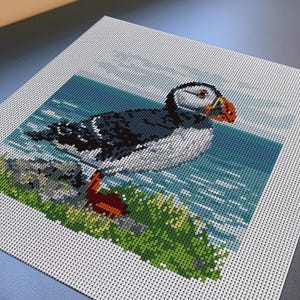Könnte beinhalten: Ein quadratisches Diamond-Painting-Set mit einem verpixelten Bild eines Papageientauchers, der auf einer grasbewachsenen Klippe steht. Der Vogel hat ein schwarz-weißes Gefieder und einen leuchtend orangefarbenen Schnabel. Der Hintergrund zeigt einen blauen Ozean.