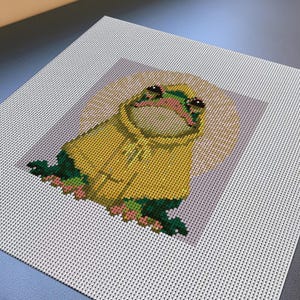 May include: A square needlepoint canvas featuring a pixelated frog wearing a yellow hooded raincoat. The frog is positioned against a circular beige background, surrounded by small green leaves and pink flowers, all rendered in a grid-like pattern.