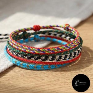 May include: A stack of four braided cord bracelets featuring various patterns and colors including red, green, blue, yellow, and black. The bracelets are constructed from thin, woven threads with a visible chevron or herringbone texture throughout.