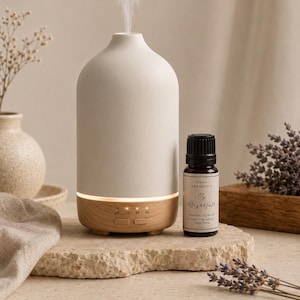 May include: A matte white ceramic essential oil diffuser with a light wood grain base and two control buttons, emitting a thin stream of mist. Beside it stands a small amber glass bottle labelled Elvan Aromatics Nightfall Essential Oil Blend.