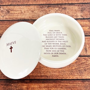 May include: A round white ceramic trinket box with a removable lid. The lid features the name MAEVE in dark red text above a small red cross. The interior base is printed with the Hail Mary prayer in dark red text, beginning with Hail Mary, full of grace, the Lord is with thee, blessed art thou amongst women, and blessed is the fruit of thy womb, Jesus. Holy Mary, Mother of God, pray for us sinners, now and at the hour of our death. Amen.