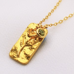 May include: A gold necklace with a rectangular pendant featuring a textured flower design. The pendant has a small green gemstone attached to it.