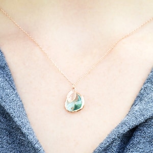 The necklace is shown on a model wearing a grey button down shirt. A rose gold metal chain with aquamarine charm and leaf charm hangs around the model's neck. The aquamarine is framed in rose gold and the leaf on top is stamped with an initial.