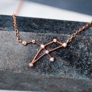 Puede incluir: Un delicado collar de oro rosa con un diseño de constelación adornado con pequeños diamantes brillantes. El collar se muestra sobre una piedra gris oscura.