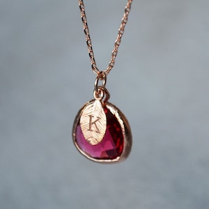 A pinkish red ruby colored transparent glass stone charm is framed in rose gold metal and hangs from a rose gold link chain. There is a small leaf shaped charm with the letter K stamped on the front, hanging in front of the ruby charm.