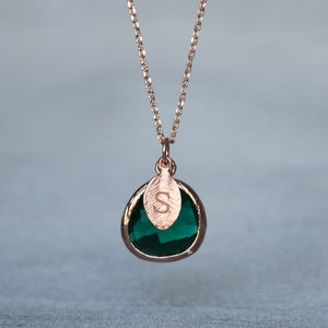 A green emerald colored transparent glass stone charm is framed in rose gold metal and hangs from a rose gold link chain. There is a small leaf shaped charm with the letter S stamped on the front, hanging in front of the emerald charm.