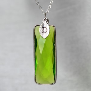 Op de afbeelding: Een zilveren ketting met een groene kristallen hanger. De hanger is rechthoekig en heeft een zilveren blaadje met de letter "D" erop gegraveerd.