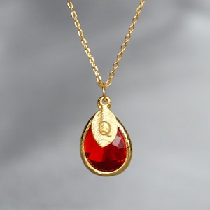 May include: A gold-colored necklace with a teardrop-shaped, red gemstone pendant. The pendant is framed in gold and features a leaf design with the letter "Q". The necklace has a delicate chain and is displayed against a soft gray background.