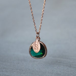 A green emerald colored transparent glass stone charm is framed in rose gold metal and hangs from a rose gold link chain. There is a small leaf shaped charm with the letter S stamped on the front, hanging in front of the emerald charm.