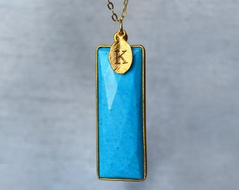 May include: A gold chain necklace with a turquoise blue rectangular pendant and a small gold charm with the letter 'K' engraved on it.