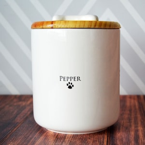 May include: White ceramic pet food container with a wooden lid. The container has the name "Pepper" and a paw print on the front.