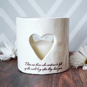 Product image of an off white ceramic votive with a white tea candle inside. The candle is visible through a heart shaped cutout on the front and back. The votive has two lines of centered light brown sepia text below the heart on the front.