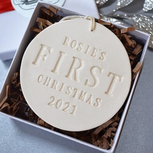 A round shaped, gloss glazed, off-white, ceramic ornament sits in a white gift box on a grey table. The ornament has a white cord to hang from and text stamped into the surface on the front. The text is in all capital letters and centered.