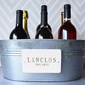 May include: A silver metal ice bucket with a white sign that reads "LANCLOS. EST. 2021."  The bucket is filled with five bottles of wine.