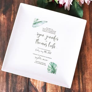 White ceramic square plate with a wedding invitation printed on it. The invitation features a tropical leaf design and the text "With great joy, William Holly Holland and Mary Elizabeth Graham invite you to celebrate the marriage of their daughter Ryan Jennifer to Thomas Lush at 4 o'clock in the evening Friday, May 19th, 2017 Ceremony on the beach of Las Palmas Sayulita, Mexico Reception to follow at Palapa Azul del Mar"