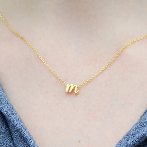 Puede incluir: Un collar de cadena dorada con un pequeño colgante de letra 'M' en cursiva.