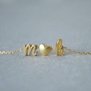 以下が含まれることがあります： ハート型のチャームで区切られた文字「m」と「h」が特徴の繊細なゴールドネックレス。チェーンは細く、ペンダントは筆記体です。ネックレスは明るい灰色の表面に表示されています。