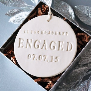 Product image of an off-white gloss-glazed ceramic round-shaped  ornament. The front is stamped with custom text in all capital letters. The text is centered and reads Alison & Johnny, Engaged, 07.07.15. The ornament has a white cord to hang from.
