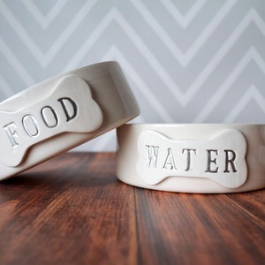 Two white ceramic dog bowls with bone-shaped tile on front. The bowl on the left says "FOOD" and the bowl on the right says "WATER".