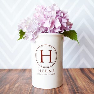 May include: A white ceramic vase with a burgundy monogram "H" and the name "HEHNS ESTABLISHED 2017" is filled with light purple hydrangea flowers and green leaves. The vase sits on a wooden surface.