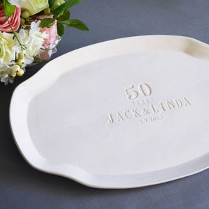 May include: An oval-shaped white ceramic serving platter featuring an embossed inscription that reads 50 YEARS JACK & LINDA 2.9.2019. The platter has a smooth, matte finish and a slightly scalloped edge, providing a clean and neutral surface.
