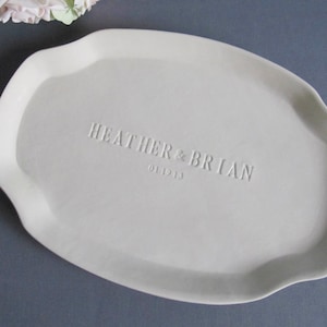 May include: A white ceramic serving platter with an irregular shape. The names "HEATHER & BRIAN" and the date "01.19.13" are embossed on the surface. The platter is displayed on a grey surface, with some flowers in the upper left corner.