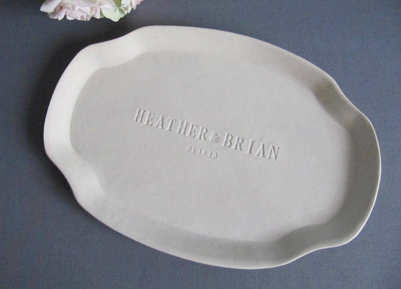 May include: A white ceramic serving platter with an irregular shape. The names "HEATHER & BRIAN" and the date "01.19.13" are embossed on the surface. The platter is displayed on a grey surface, with some flowers in the upper left corner.