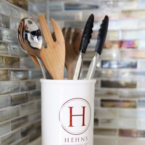 A white ceramic utensil holder with the letter H in a circle and the text 'Hehns Established 2017'. The holder is filled with kitchen utensils including a wooden spoon, a silver serving spoon, and two black tongs.