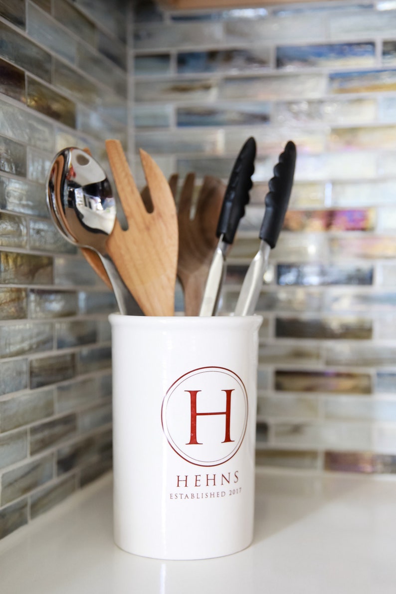 A white ceramic utensil holder with the letter H in a circle and the text 'Hehns Established 2017'. The holder is filled with kitchen utensils including a wooden spoon, a silver serving spoon, and two black tongs.