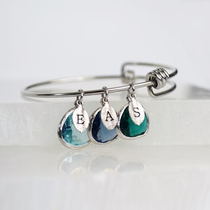 Może przedstawiać: Srebrna bransoletka z trzema zawieszkami w kształcie łzy. Zawieszki są niebieskie, turkusowe i zielone i mają wygrawerowane litery E, A i S.