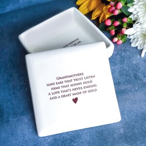 Op de afbeelding: Witte keramische doos met een deksel, met een citaat over grootmoeders: "Grootmoeders hebben oren die echt luisteren, armen die altijd vasthouden, een liefde die nooit eindigt en een hart van goud." Het citaat is gedrukt in kastanjebruine inkt met een klein rood hart aan het einde.