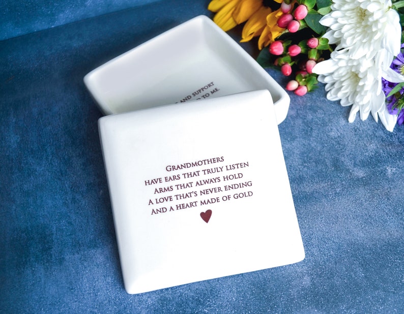 Op de afbeelding: Witte keramische doos met een deksel, met een citaat over grootmoeders: "Grootmoeders hebben oren die echt luisteren, armen die altijd vasthouden, een liefde die nooit eindigt en een hart van goud." Het citaat is gedrukt in kastanjebruine inkt met een klein rood hart aan het einde.