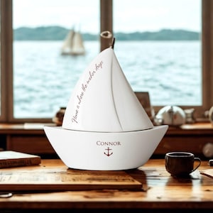 ceramic sailboat shaped keepsake box on a wood desk in front of a window with the ocean in background. Box is personalized as a gift