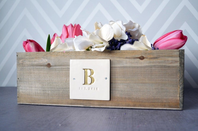 A wooden planter box with a white ceramic plaque personalized with initial and date