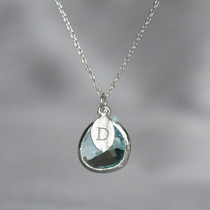 A light blue aquamarine colored transparent glass stone charm is framed in silver metal and hangs from a silver link chain. There is a small leaf shaped charm with the letter D stamped on the front, hanging in front of the aquamarine charm.