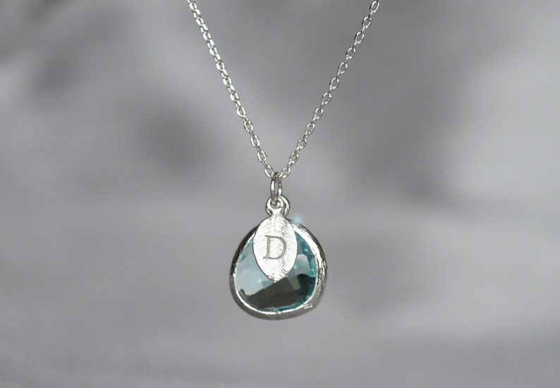 A light blue aquamarine colored transparent glass stone charm is framed in silver metal and hangs from a silver link chain. There is a small leaf shaped charm with the letter D stamped on the front, hanging in front of the aquamarine charm.