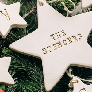 Five white ceramic star ornaments with gold lettering. The largest star has the text 'THE SPENCERS' on it. The other stars have the letters 'V', 'S', and 'S' on them. The ornaments are surrounded by green pine branches.