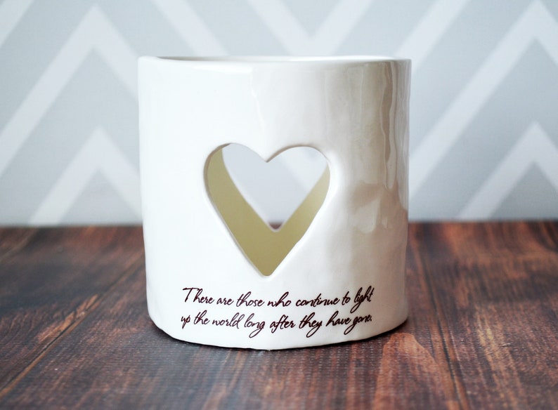 Product image of an off white ceramic votive. You can see through the votive through a heart shaped cutout on the front and back. The votive has two lines of centered light brown sepia text below the heart on the front.