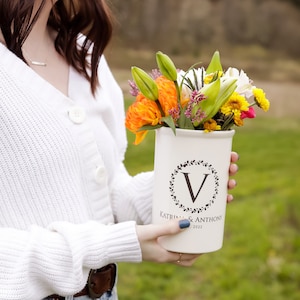 May include: A white ceramic vase with a black wreath and the letter "V" in the center. The vase has the text "Katrina & Anthony 2022" printed on the bottom. The vase is filled with a bouquet of orange, yellow, and white flowers.