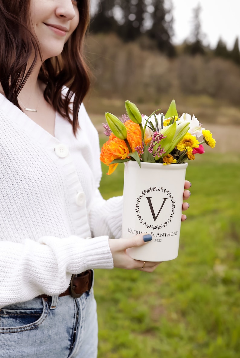 May include: A white ceramic vase with a black wreath and the letter "V" in the center. The vase has the text "Katrina & Anthony 2022" printed on the bottom. The vase is filled with a bouquet of orange, yellow, and white flowers.