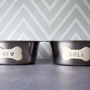 May include: Two stainless steel pet bowls with bone-shaped name tags. One tag reads "WE ❤️" and the other reads "LOLA". The bowls are set against a gray and white chevron patterned background.
