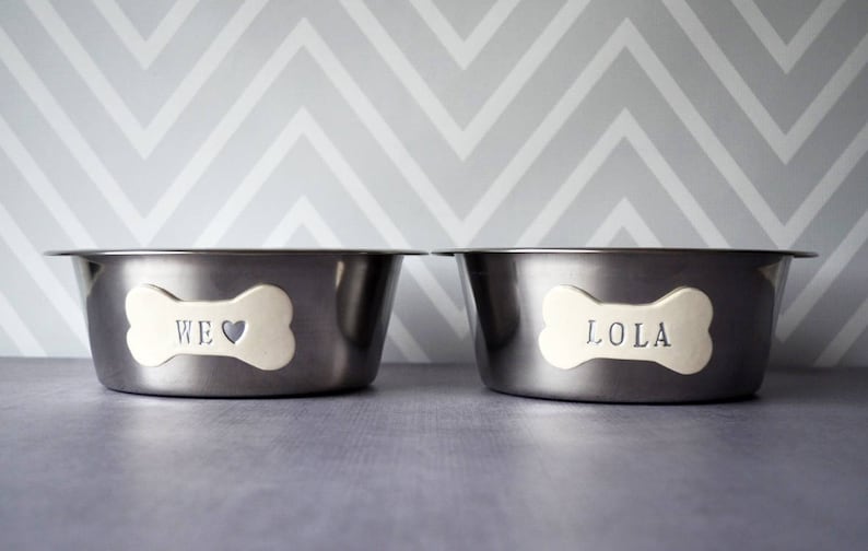May include: Two stainless steel pet bowls with bone-shaped name tags. One tag reads "WE ❤️" and the other reads "LOLA". The bowls are set against a gray and white chevron patterned background.