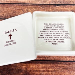 Könnte beinhalten: Weiße Keramikbox mit einem roten Kreuz und dem Namen Isabella. Die Box ist mit dem Text "Getauft am 10. Juni 2025" beschriftet. Der Deckel der Box ist mit einem spanischen Gebet beschriftet.