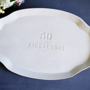 Off-white ceramic serving platter with an irregular, wavy edge. The platter is embossed with the text "50 YEARS JACK & LINDA 2.9.2013". A floral arrangement is visible in the upper left corner.