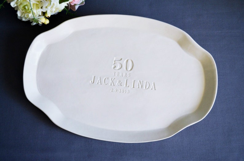 Off-white ceramic serving platter with an irregular, wavy edge. The platter is embossed with the text "50 YEARS JACK & LINDA 2.9.2013". A floral arrangement is visible in the upper left corner.