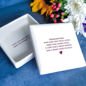 Op de afbeelding: Witte keramische doos met deksel. Het deksel heeft de tekst "Your love and support mean the world to" erop gedrukt. De doos heeft de tekst "Grandmothers have ears that truly listen, arms that always hold, a love that's never ending and a heart made of gold" erop gedrukt. De tekst is in een kastanjebruine kleur. Een klein rood hartje is onder de tekst gedrukt.