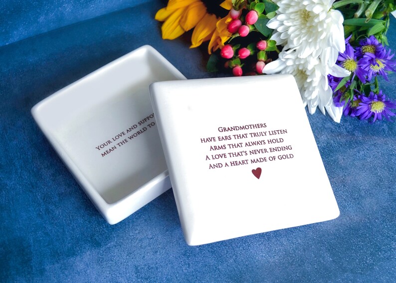 Op de afbeelding: Witte keramische doos met deksel. Het deksel heeft de tekst "Your love and support mean the world to" erop gedrukt. De doos heeft de tekst "Grandmothers have ears that truly listen, arms that always hold, a love that's never ending and a heart made of gold" erop gedrukt. De tekst is in een kastanjebruine kleur. Een klein rood hartje is onder de tekst gedrukt.