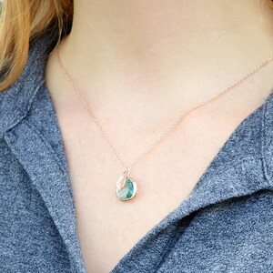 May include: Rose gold necklace with a pendant featuring a teal and white stone. The pendant is round with a leaf-like design. The necklace is worn against a gray shirt.