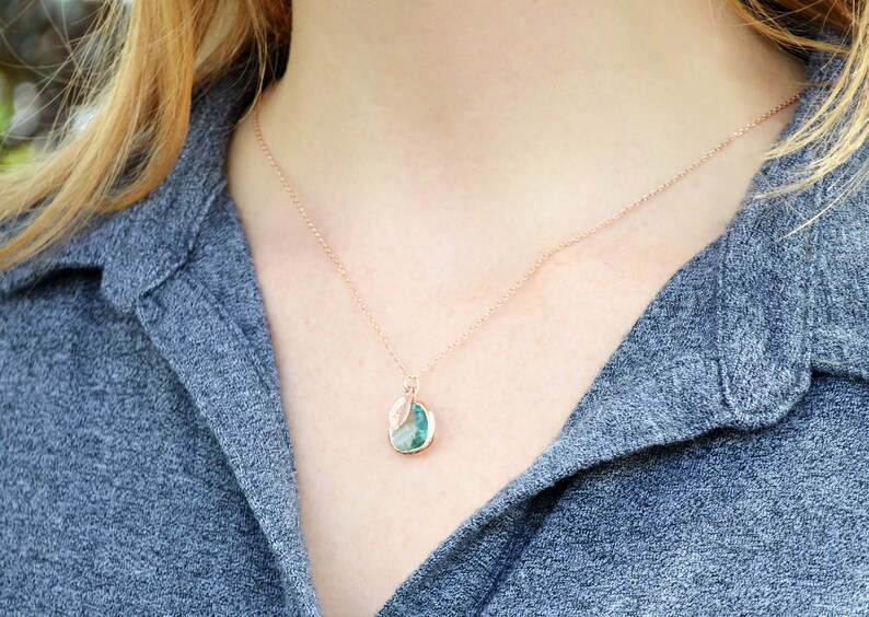 May include: Rose gold necklace with a pendant featuring a teal and white stone. The pendant is round with a leaf-like design. The necklace is worn against a gray shirt.