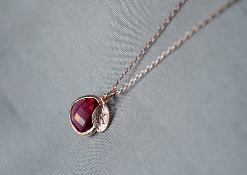 A pinkish red ruby colored transparent glass stone charm is framed in rose gold metal and hangs from a rose gold link chain. There is a small leaf shaped charm with the letter K stamped on the front, hanging in front of the ruby charm.